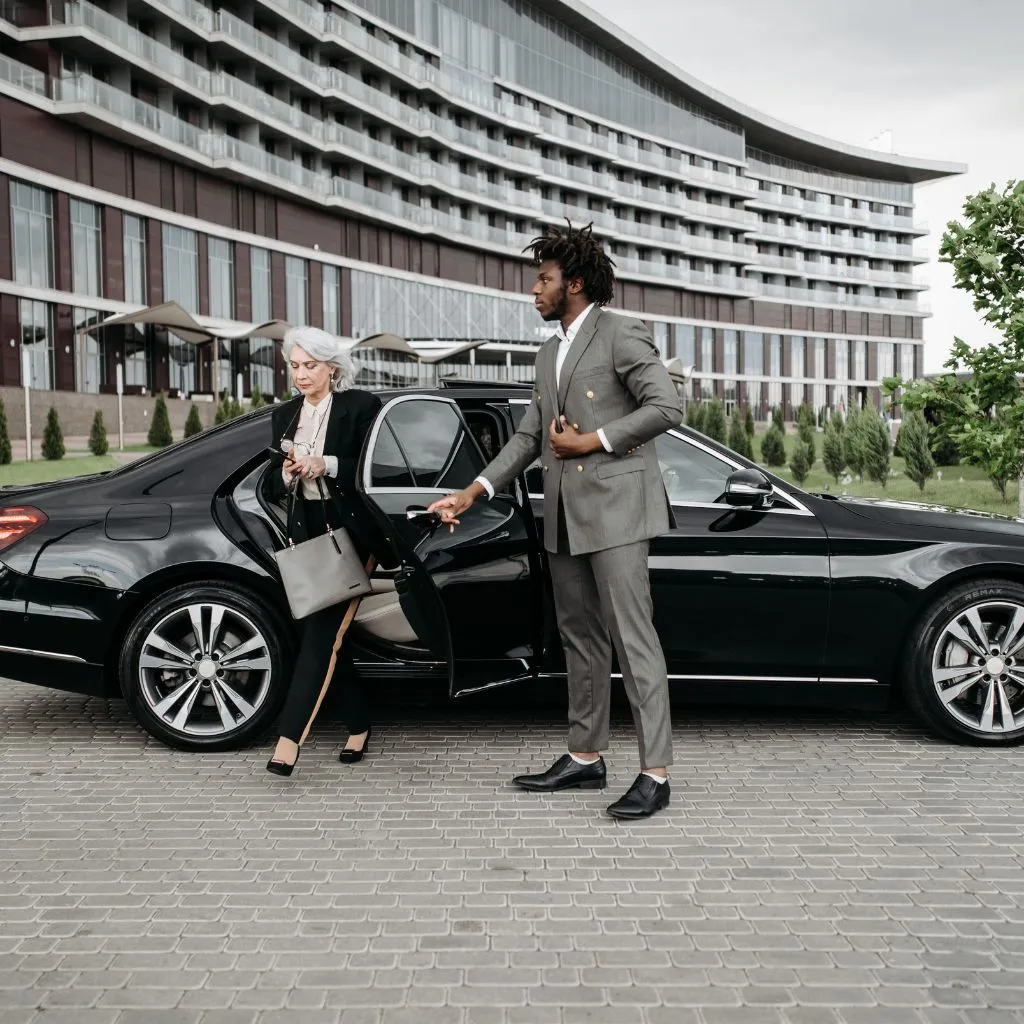 A professional, well-dressed chauffeur in a grey suit assists a senior businesswoman as she exits a sleek black luxury sedan (executive car) outside a modern hotel or corporate building. This depicts premium Barrie Limo Service for corporate transportation or airport transfers.