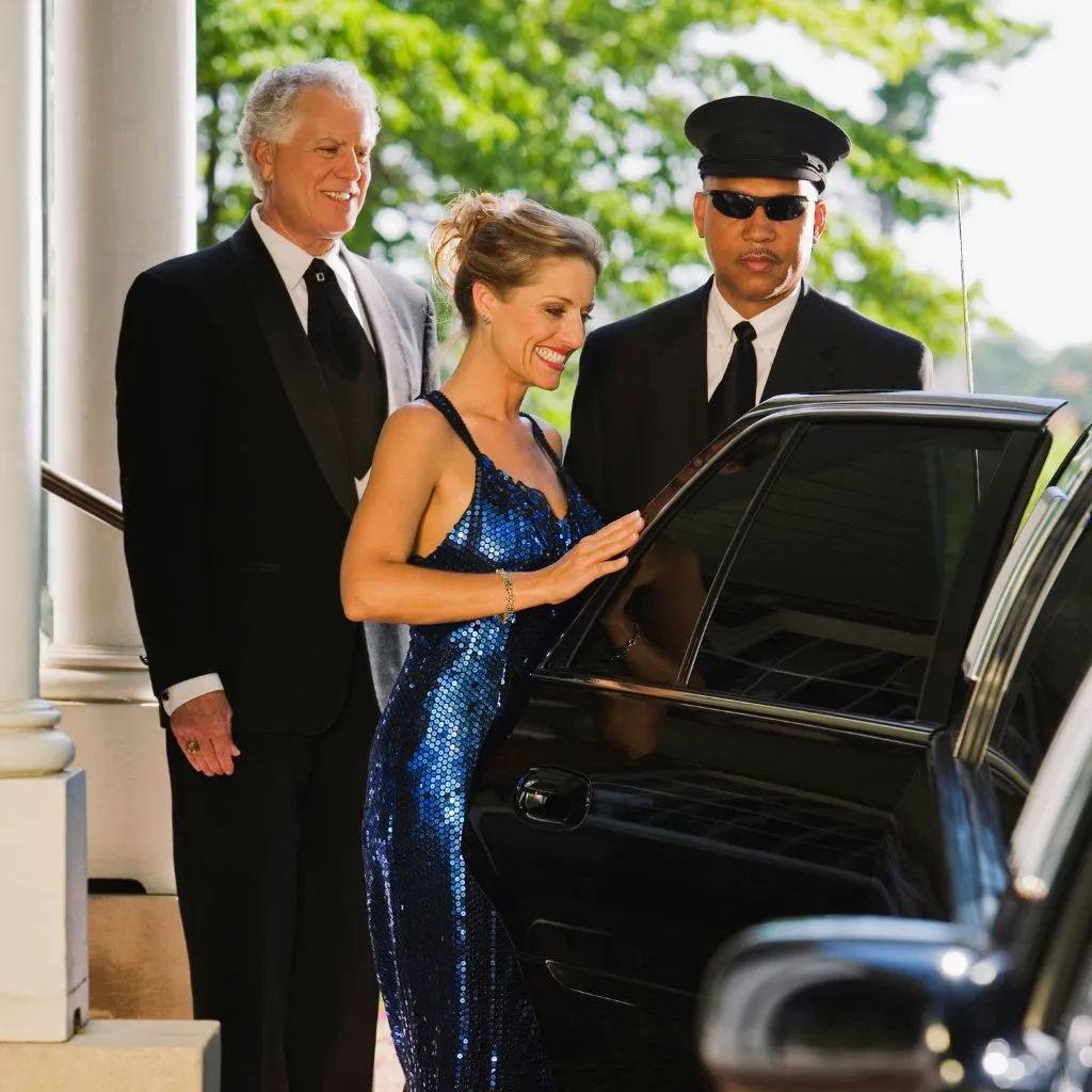 A glamorous woman in a blue sequined gown is being assisted by a professional chauffeur as she steps into a black limousine. A well-dressed man in a tuxedo stands beside her. The image suggests a luxury transportation experience, like an Orillia Limousine Service.